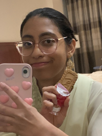 A selfie of Batool Kazmi smiling while holding her phone and a cookie. She is wearing a white attire, with big golden earings.